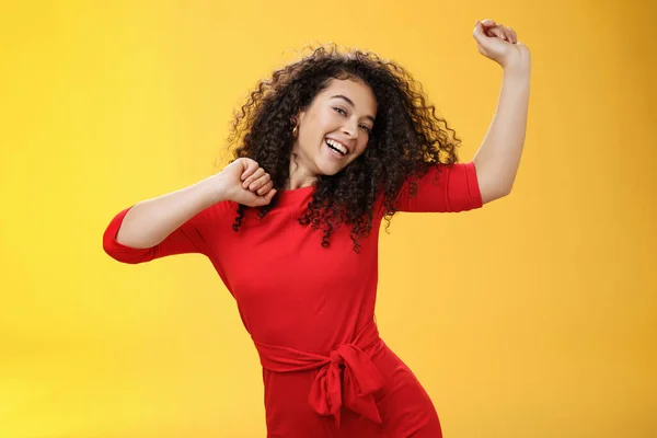 Zorgeloos meisje voelt zich verheven en vrolijk dansen in rode jurk handen opheffen vrolijk kantelen hoofd en breed glimlachen op de camera als genieten van vakantie, feestdagen te vieren over gele achtergrond — Stockfoto