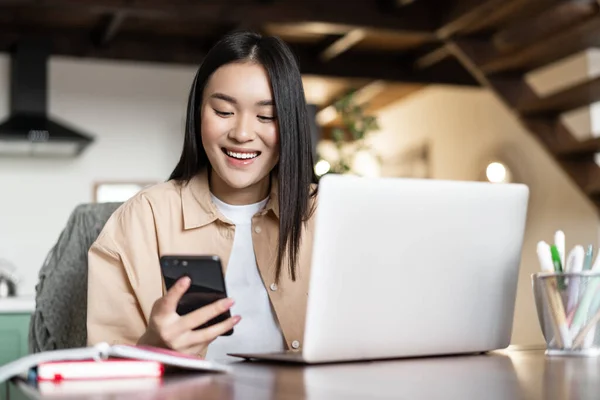 Sonriente asiática chica estudiante haciendo la tarea en el portátil. Mujer usando el teléfono celular y la computadora para trabajar desde casa — Foto de Stock