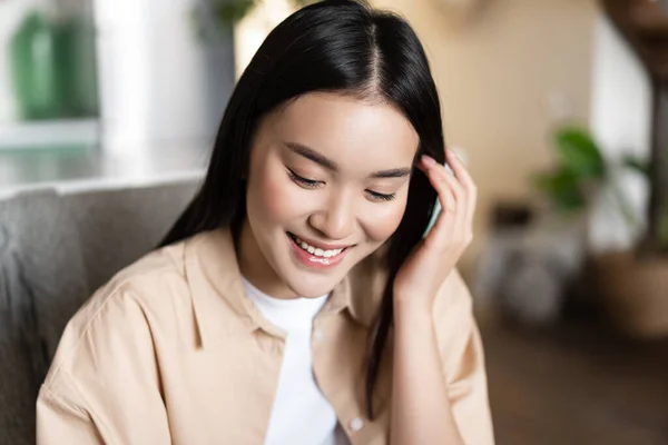 Sevimli Asyalı kız öğrenci gülümsüyor, aşağı bakıyor ve saçlarını kulaklarının arkasına sokuyor, evde günlük kıyafetlerle oturuyor. — Stok fotoğraf