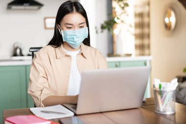 Medikal maskeli Asyalı bir kız evde çalışıyor. Morali bozukken, dizüstü bilgisayar kullanıyor.