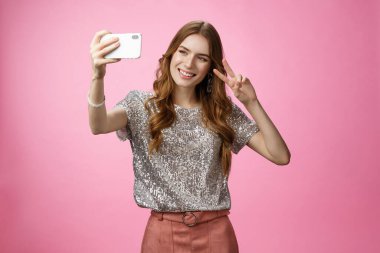 Çekici, çekici, beyaz bir kadın selfie çekiyor. Zafer işareti, gülümseyen, akıllı telefon, sırıtan cep telefonu görüntüsü, video çağrısı, pembe arka plan.