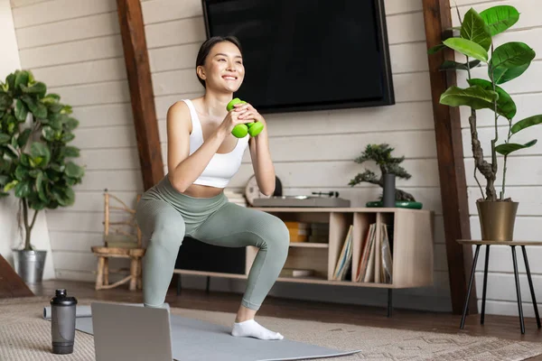 Young asian woman doing workout at home with dumbbels, making squats on floor mat, follow online fitness instructor guide on laptop — Stock Photo, Image