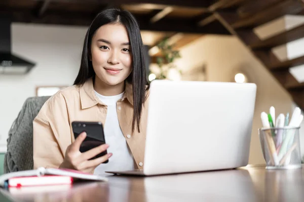 Sonriendo chica asiática utiliza la computadora en casa, sostiene el teléfono inteligente y se ve feliz en la cámara. Mujer ocupada trabajando a distancia en el ordenador portátil — Foto de Stock