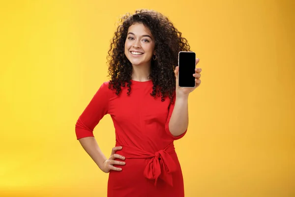Flicka skryta med ny telefon hon fick på jul känsla glad hålla mobil enhet i handen visar smartphone skärm t kamera, ler brett med upplyft humör över gul bakgrund — Stockfoto