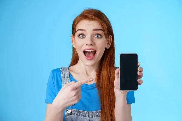 Eccitato divertito bella ragazza rossiccio promuovere app impressionante, visualizzare display smartphone, puntando schermo del telefono stupito, sorridente sorpreso, come il gioco fresco, stand sfondo blu — Foto Stock