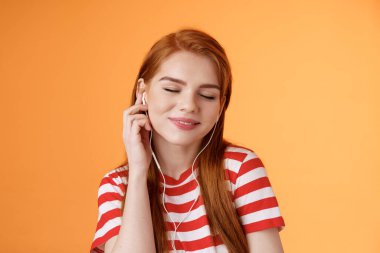 Güzel, çekici, kızıl saçlı, 20 'li yaşlarda bir kadın müzik dinler, gözleri kapar, gülümser, kulaklığa dokunur, kafa sallar, keyif alır, güzel ritimler dinler, en sevdiği müzik parçası, turuncu arka plan.