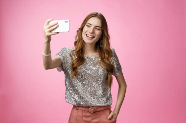 Sosyal görünüşlü, kendine güvenen, beyaz bir kadın video mesajı kaydediyor. Elinde akıllı telefon, elinde resim partileri, internette pembe arka plan pozu veriyor.