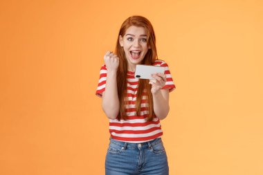Heyecanlı kızıl saçlı kadın pas düzeyi, harika bir oyun gibi, gol, akıllı telefonu yatay tut, yumruk pompası başarısı, şanslı başarıyı kutla, geniş gülümse, turuncu arka plan