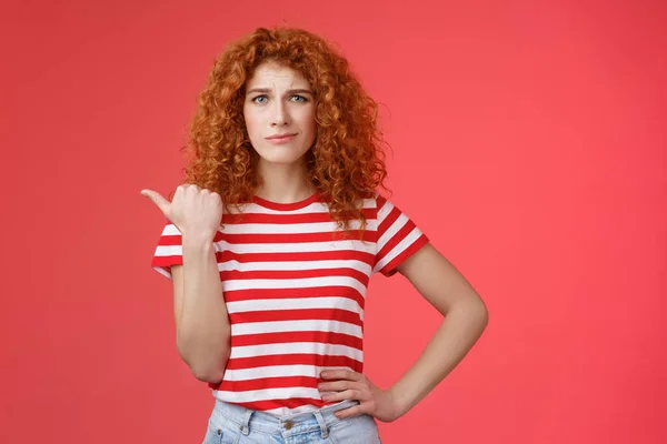 Girl not sure best choice. Doubtful redhead curly woman look suspicious frowning uncertain stare hesitant pointing left thumb unsure buy or not standing red background displeased — Stock Photo, Image