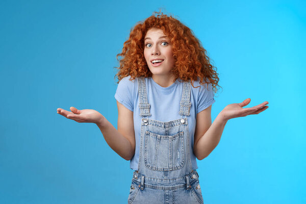 Who knows. Careless clueless attractive playful redhead cute curly-haired girl shrugging unaware hold hands sideways unaware smiling act innocent uninvolved not knwo answer blue background