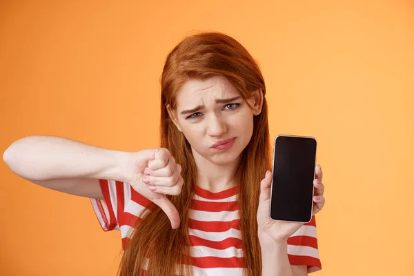 stock image Very bad poor quality. Upset redhead female customer grimace disappointed, show smartphone blank display, thumbs down judgement, dislike app, smirk dismay, stand upset orange background