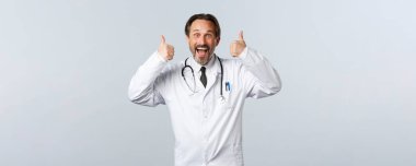 Covid-19, koronavirüs salgını, sağlık çalışanları ve pandemik konsept. Heyecanlı, neşeli, beyaz önlüklü erkek doktor onayladı. Cerrah kabul ediyor ya da fikir gibi, tanıtım öneriyor.