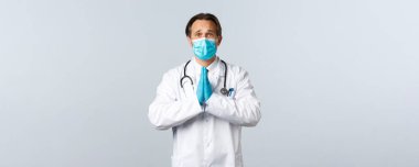 Covid-19, virüsü önleme, sağlık çalışanları ve aşı konsepti. Umut dolu, tıbbi maskeli ve eldivenli doktor. Dua ederken, kaşlarını çatarken, iyilik isterken.