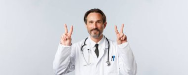 Covid-19, virüsü önleme, sağlık çalışanları ve aşı konsepti. Beyaz önlüklü neşeli, orta yaşlı doktor barış işaretleri gösteriyor ya da V işareti ve gülümsüyor. Olumlu düşün.