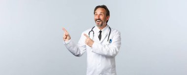 Covid-19, koronavirüs salgını, sağlık çalışanları ve pandemik konsept. Beyaz önlüklü, gülümseyen erkek doktor sol üst köşesini işaret ederek eğleniyor, ilaç veya klinik afişi gösteriyor.