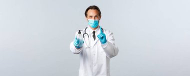 Covid-19, virüsü önleme, sağlık çalışanları ve aşı konsepti. Hevesli gülümseyen doktor el dezenfektanı kullanmanın önemini açıklar, birinci kuralı gösterir, tıbbi maske takar ve eldiven giyer.