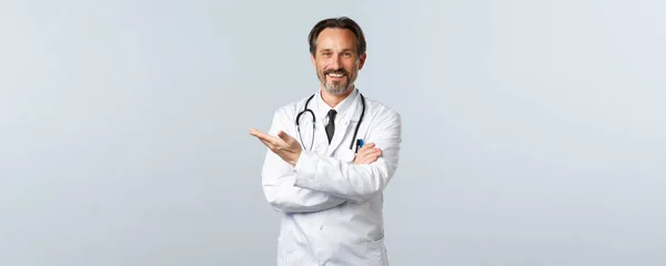 Covid-19, koronavirüs salgını, sağlık çalışanları ve pandemik konsept. Yakışıklı dost canlısı doktor yolu gösteriyor. Beyaz önlüklü doktor tanıtımda sol elini göstererek hastaları davet ediyor. — Stok fotoğraf