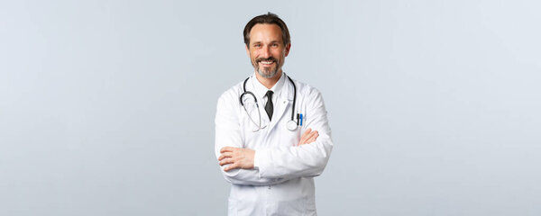 Ковид-19, вспышка коронавируса, медицинские работники и пандемия. Энтузиастичный улыбающийся доктор, врач в белом халате, с энтузиазмом смотрящий, скрещенные руки груди, слушающий пациента