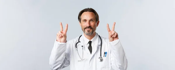Covid-19, Prävention von Viren, Gesundheitspersonal und Impfkonzept. Gut aussehender Arzt mittleren Alters in weißem Mantel, der Friedenszeichen oder V-Zeichen zeigt und lächelt, bleibt positiv — Stockfoto