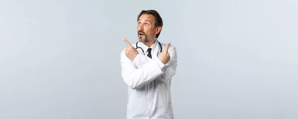 Covid-19, coronavirus outbreak, healthcare workers and pandemic concept. Unsure indecisive doctor in white coat, pointing fingers sideways and look left at variant, making choice — Stock Photo, Image