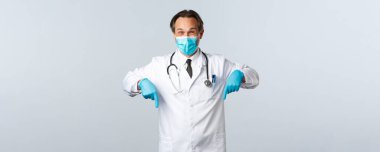 Covid-19, virüsü önleme, sağlık çalışanları ve aşı konsepti. Hevesli, yakışıklı, orta sınıf maskeli ve eldivenli bir erkek doktor. İlanı işaret ediyor. Beyaz arka plan.