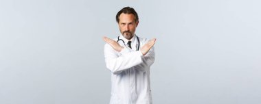 Covid-19, koronavirüs salgını, sağlık çalışanları ve pandemik konsept. Beyaz önlüklü ciddi bir erkek doktor, el ele tutuşup jesti durduruyor, yasaklıyor ya da yasaklıyor.
