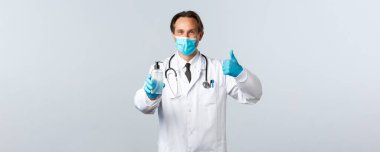 Covid-19, virüsü önleme, sağlık çalışanları ve aşı konsepti. Tıbbi maskeli tatmin olmuş bir doktor ve onaylı eldivenler, el dezenfektanı, beyaz arka plan.