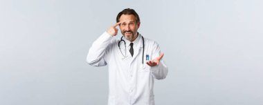 Covid-19, koronavirüs salgını, sağlık çalışanları ve pandemik konsept. Üzgün erkek doktor hasta azarlıyor aptalca davranıyor, tapınağı işaret ediyor ve umutsuzca elini kaldırıyor, yüz yüze hayal kırıklığına uğruyor.