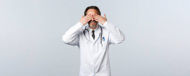 Covid-19, koronavirüs salgını, sağlık çalışanları ve pandemik konsept. Şaşırmış beyaz önlüklü mutlu erkek doktor elleri kapalı ve bekliyor, sürpriz bekleniyor, beyaz arka plan
