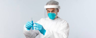 Covid-19, virüsü önleme, sağlık çalışanları ve aşı konsepti. Ciddi görünümlü bir doktor, klinikte teknoloji laboratuarı koruyucu ekipman giyiyor, test tüpünü mavi sıvıyla tutuyor.