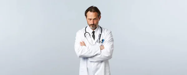 Covid-19, coronavirus outbreak, 의료 종사자와 전염병 컨셉트. 심각하게 실망 한 흰 코트를 입은 남자 의사, 양팔 가슴을 교차하는 모습 그리고 이마의 판 단 아래서 바라봄 — 스톡 사진