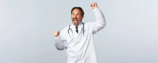 Covid-19, ξέσπασμα του ιού της στέψης, επαγγελματίες υγείας και πανδημία. Ενθουσιασμένος άντρας γιατρός με λευκό παλτό σηκώνει το χέρι και φωνάζει ναι, βλέποντας αθλητικό αγώνα, νικώντας, θριαμβεύοντας. — Φωτογραφία Αρχείου