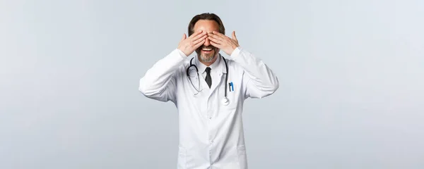 Covid-19, Coronavirus-Ausbruch, Gesundheitspersonal und Pandemiekonzept. Überrascht glücklicher Arzt im weißen Mantel schließt die Augen mit den Händen und wartet, antizipiert Überraschung, weißer Hintergrund — Stockfoto