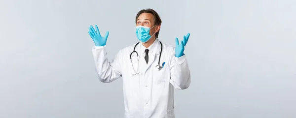 Covid-19, πρόληψη του ιού, εργαζόμενοι στον τομέα της υγείας και την έννοια του εμβολιασμού. Ενθουσιασμένος και ανακουφισμένος άνδρας γιατρός με ιατρική μάσκα, γάντια, σηκώνοντας τα χέρια ψηλά χαρούμενος, κοιτάζοντας πάνω αριστερή γωνία ευχαριστημένος — Φωτογραφία Αρχείου