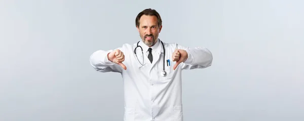 Covid-19, ξέσπασμα του ιού της στέψης, επαγγελματίες υγείας και πανδημία. Θυμωμένος απογοητευμένος άντρας γιατρός με λευκό παλτό, παραπονιέται για έντονη απέχθεια, δείχνει τους αντίχειρες προς τα κάτω — Φωτογραφία Αρχείου
