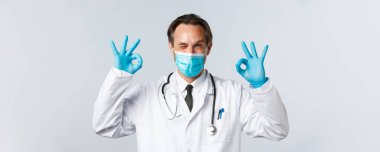 Covid-19, virüsü önleme, sağlık çalışanları ve aşı konsepti. Tıbbi maskeli ve eldivenli dost canlısı gülümseyen doktor, göz kırp ve iyi niyet göster, garanti et ya da hizmet et.