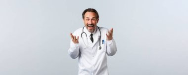 Covid-19, koronavirüs salgını, sağlık çalışanları ve pandemik konsept. Heyecanlı mutlu erkek doktor gülüyor ve komik bir şeyi açıklıyor, geniş bir gülümseme, beyaz arka plan