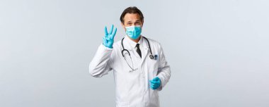 Covid-19, virüsü önleme, sağlık çalışanları ve aşı konsepti. Tıbbi maskeli ciddi bir doktor ve eldivenler sosyal mesafeyi açıklar, üç parmak göster, beyaz arka plan