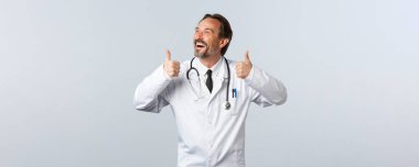 Covid-19, koronavirüs salgını, sağlık çalışanları ve pandemik konsept. Beyaz önlüklü kaygısız, mutlu erkek doktor, sol üst köşeye bakıyor ve kahkahalar atıyor.