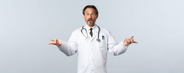 Covid-19, koronavirüs salgını, sağlık çalışanları ve pandemik konsept. Beyaz önlüklü, tereddütlü erkek doktor, elleri yana doğru uzanıyor ve kafası karışık bakıyor, karar veremiyorum.
