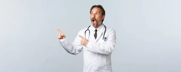 Covid-19, coronavirus outbreak, 의료 종사자와 전염병 컨셉트. 흰색 외투를 입고 손가락을 왼쪽 위로 하고 광고에 턱을 떨어뜨리는 깜짝 놀란 말 못 하는 의사, 프로 모를 보여 주는 — 스톡 사진