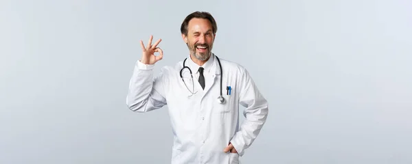 Covid-19, coronavirus outbreak, healthcare workers and pandemic concept. Cheerful happy doctor in white coat assure or recommend medicine, showing okay guarantee gesture and wink — Stock Photo, Image