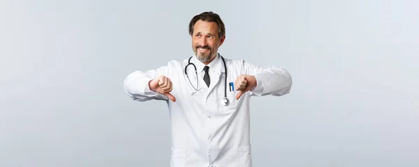 Covid-19, ξέσπασμα του ιού της στέψης, επαγγελματίες υγείας και πανδημία. Σκεπτικός και απρόθυμος άντρας γιατρός με λευκό παλτό κοιτάζει μακριά γκρινιάζοντας δυσαρεστημένος, δείχνει τους αντίχειρες προς τα κάτω — Φωτογραφία Αρχείου