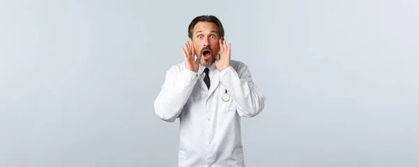 Covid-19, coronavirus outbreak, healthcare workers and pandemic concept. Shocked excited male doctor in white coat calling someone, shouting loud with hands near opened mouth — Stock Photo, Image