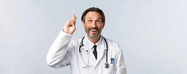 Covid-19, zapobieganie wirusom, pracownikom służby zdrowia i koncepcji szczepień. Zbliżenie optymistycznego, pełnego nadziei lekarza w białym płaszczu, trzymając kciuki za szczęście i patrząc w górę uśmiechając się, wypowiadając życzenie — Zdjęcie stockowe