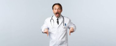 Covid-19, koronavirüs salgını, sağlık çalışanları ve pandemik konsept. Beyaz önlüklü şaşkın doktor, çenesini indir ve reklama işaret et, tanıtımın yolunu göster