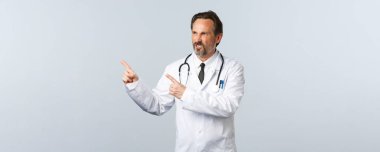 Covid-19, koronavirüs salgını, sağlık çalışanları ve pandemik konsept. Sinirli doktor küfrediyor, sol üst köşeyi işaret ediyor ve kaşlarını çatıyor, şikayet ediyor.