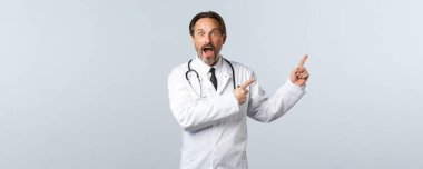 Covid-19, koronavirüs salgını, sağlık çalışanları ve pandemik konsept. Etkileyici ve heyecanlı beyaz önlüklü erkek doktor, harika haberlere tepki veriyor, sağ üst köşeyi işaret ediyor.