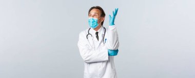 Covid-19, virüsü önleme, sağlık çalışanları ve aşı konsepti. Heyecanlı, sağlık maskeli ve eldivenli erkek doktor, düşünceli el kaldırıyor, bir fikri var, şaşırmış görünüyor.