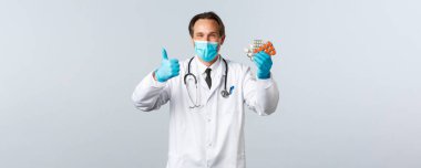 Covid-19, virüsü önleme, sağlık çalışanları ve aşı konsepti. Tatmin olmuş yakışıklı doktor gülümsüyor, tıbbi maske ve eldiven takıyor, tavsiye hapları olarak baş parmağını kaldırıyor, iyi ilaçlar gösteriyor.